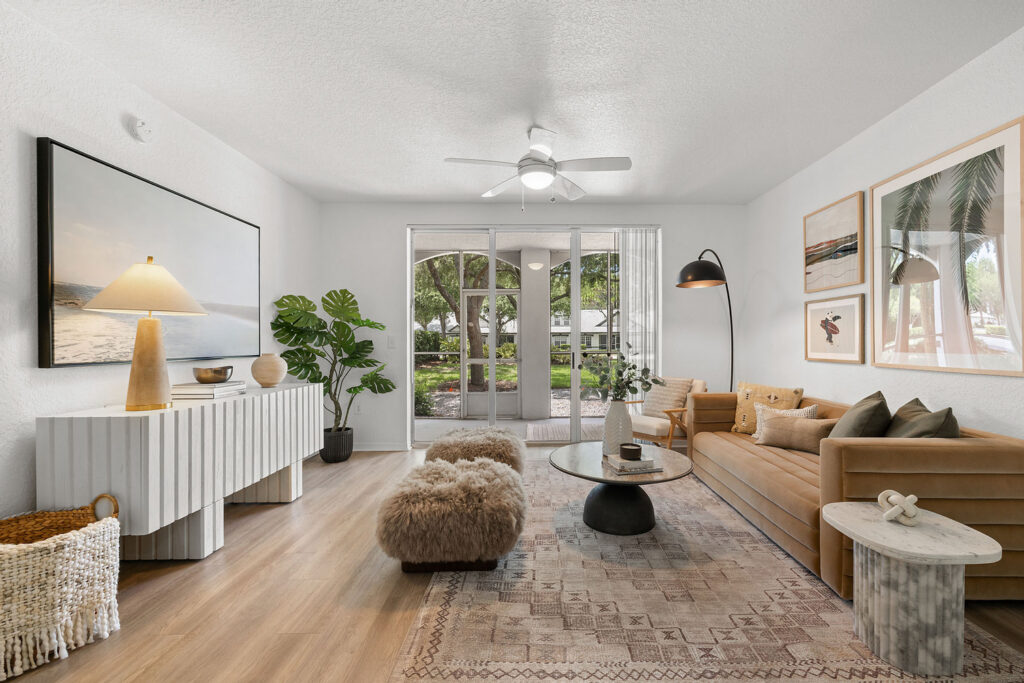 Modern living room with tan sofa, round coffee table, two fuzzy ottomans, and decorative art on the walls, featuring large windows and a view of greenery outside. Light wood floors and neutral decor create a bright, cozy space.