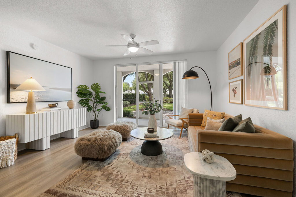 Modern living room with a tan sofa, two furry ottomans, a round coffee table, potted plant, wall art, and large windows overlooking a green outdoor area. Neutral tones and cozy, stylish decor.