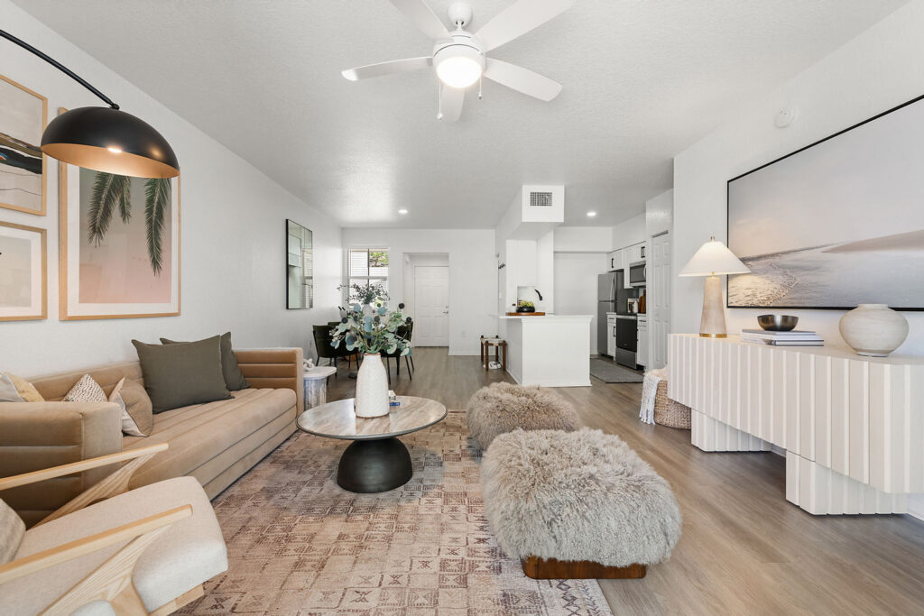 A modern living room with neutral tones features a beige sofa, round coffee table, two furry stools, and wall art. The space connects to an open kitchen with white cabinetry and stainless steel appliances.