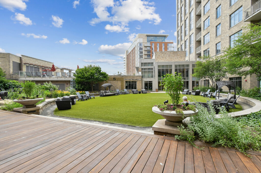 A landscaped courtyard with artificial grass, outdoor seating, and garden planters, surrounded by modern apartment buildings under a blue sky with scattered clouds.